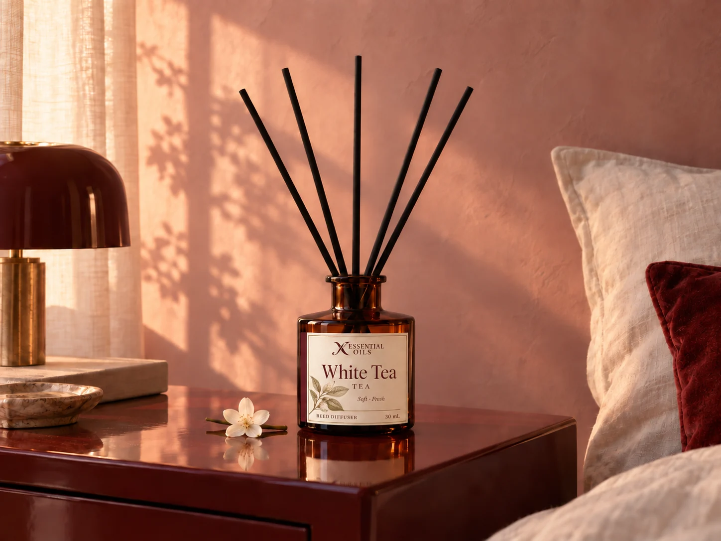 An amber reed diffuser with five black reeds on an oxblood bedside cabinet in warm morning light